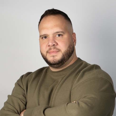 Professional portrait of a person with short dark hair and a beard, wearing an olive green sweatshirt. They have their arms crossed and are looking directly at the camera. The photo is taken against a light gray background. 